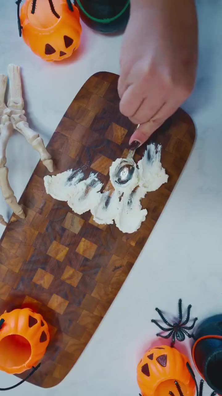 Mano untando queso crema en forma de fantasma sobre una tabla con decoración de Halloween, vista cenital